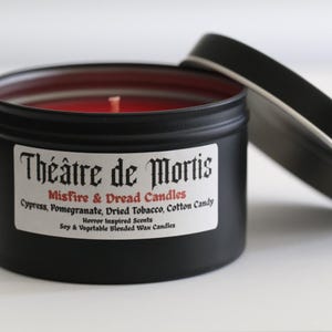 May include: A black candle tin with a red wax interior and a partially removed lid. The label reads "Théâtre de Mortis" and lists scent notes like cypress and pomegranate. The candle is a horror-inspired scent made with soy and vegetable wax.