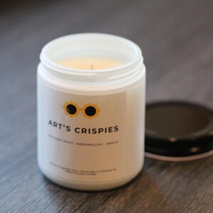 May include: A white glass candle jar with a black lid placed to the side. The label features a yellow sunflower-shaped icon with black centres, the text ART'S CRISPIES, and smaller text reading RICE CRISPY TREATS - MARSHMALLOW - VANILLA.