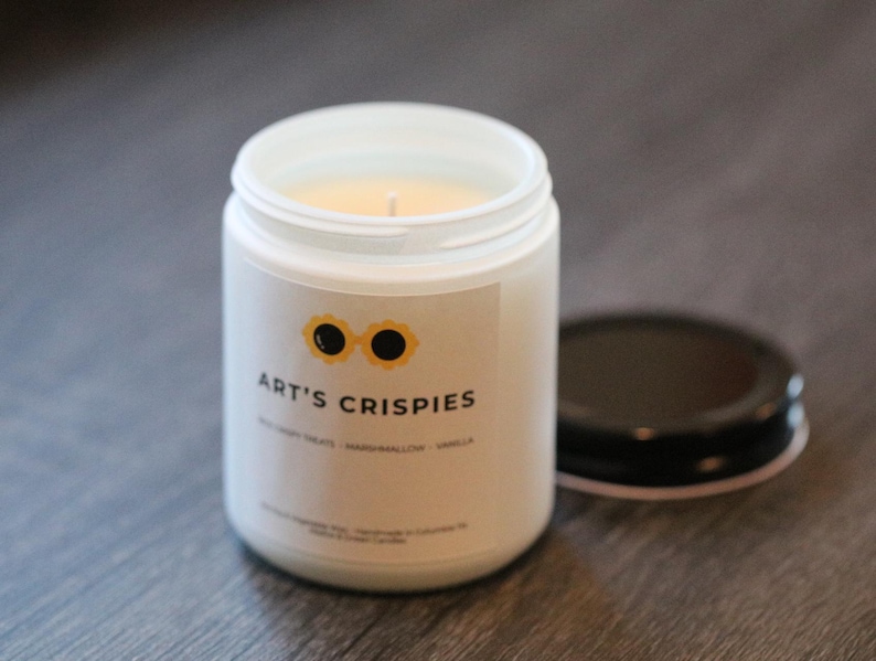 May include: A white glass candle jar with a black lid placed to the side. The label features a yellow sunflower-shaped icon with black centres, the text ART'S CRISPIES, and smaller text reading RICE CRISPY TREATS - MARSHMALLOW - VANILLA.