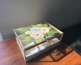 May include: A rectangular glass jewelry box with a gold-coloured metal frame. The lid is decorated with pressed flowers in shades of pink, white, and blue, with green foliage. The box is open, revealing a white interior with compartments.