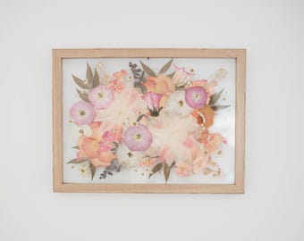 May include: A framed pressed flower arrangement with pink, peach, and white flowers. The flowers are arranged in a bouquet shape and are pressed between two pieces of glass.