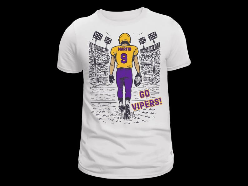 Pu&ograve; includere: Una t-shirt bianca con un'illustrazione grafica di un giocatore di football americano in maglia gialla con il nome MARTIN e il numero 9 in viola, pantaloni viola, che cammina su un campo da stadio con la scritta GO VIPERS! stampata in viola.