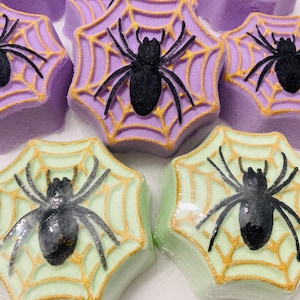 May include: Close-up of several Halloween-themed bath bombs. The bath bombs are shaped like octagons with a spider web design. The webs are gold, and the spiders are black. The bath bombs are in shades of purple and green.