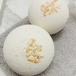 May include: Two white bath bombs with a design of oatmeal flakes on top. The bath bombs are on a gray towel.