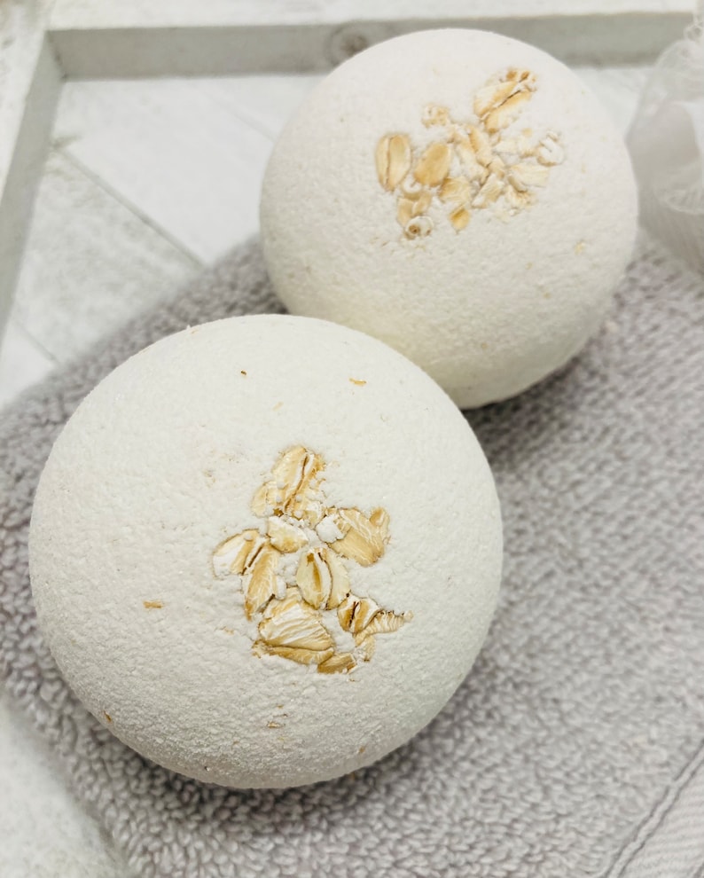 May include: Two white bath bombs with a design of oatmeal flakes on top. The bath bombs are on a gray towel.
