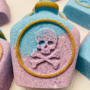 May include: A close-up of a bath bomb shaped like a potion bottle, with a skull and crossbones design. The bath bomb is a mix of blue and purple with gold glitter accents. Other bath bombs are in the background.