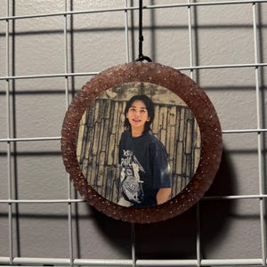 May include: A circular hanging ornament featuring a photograph of a person with shoulder-length dark hair wearing a dark graphic t-shirt. The photo is encased in a textured, translucent, light pink resin frame with a black cord for hanging.