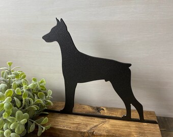 May include: Black metal silhouette of a Doberman Pinscher dog standing on a rustic wooden base.