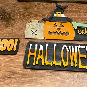 May include: Halloween decorations with a black and orange "BOO!" sign, a black sign with the word "HALLOWEEN" in orange and white, a pumpkin with a witch's hat, a poison bottle, and a cauldron with a bone and the word "eek!"