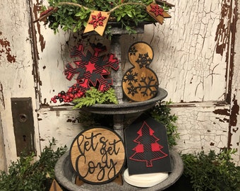 May include: A three-tiered wooden stand decorated with a red and black buffalo plaid pattern and winter-themed wooden cutouts. The stand features a deer, snowflakes, a snowman, a Christmas tree, and a sign that reads "Let's get cozy".