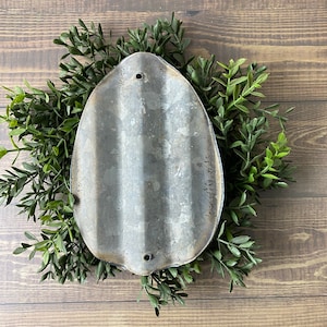 May include: A rustic, oval-shaped metal sign with a corrugated texture and two holes at the top. The sign is surrounded by a wreath of green foliage.