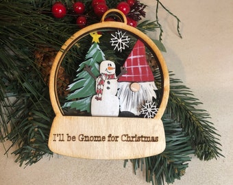 May include: Wooden Christmas ornament shaped like a snow globe with a gnome and snowman inside. The ornament has the text "I'll be Gnome for Christmas" on the bottom.