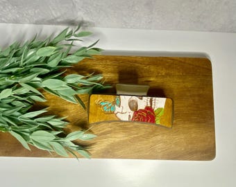 May include: A wooden hair clip with a skull and rose design. The skull is white with black details and the rose is red with green leaves. A blue butterfly is on the skull. The clip is on a wooden surface.