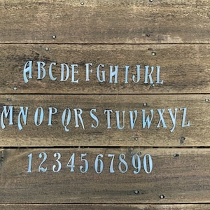 May include: A wooden surface with the alphabet and numbers 1 through 9 and 0 painted in white. The letters are in a stylized font.