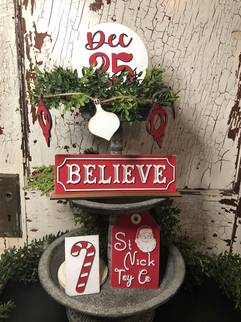 K&ouml;nnte beinhalten: Ein dreist&ouml;ckiger Holzst&auml;nder, der mit weihnachtlichen Ornamenten dekoriert ist. Die oberste Ebene zeigt einen wei&szlig;en Kreis mit dem Text "Dec 25" in Rot. Die mittlere Ebene hat ein rotes Schild mit dem Wort "BELIEVE" in Wei&szlig;. Die unterste Ebene hat ein rotes Etikett mit dem Text "St Nick Toy Co" und einem Cartoon-Bild vom Weihnachtsmann. Es gibt auch ein wei&szlig;es Zuckerstangen-Ornament.