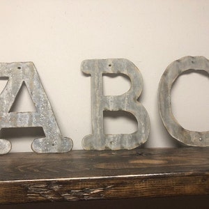 May include: Three metal letters, A, B, and C, painted in a rustic, galvanized finish. The letters are arranged in a row on a dark wood shelf.