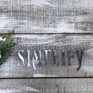 May include: Metal wall decor with the word "SIMPLIFY" in a stylized font on a whitewashed wood background.