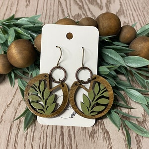 May include: A pair of green and brown wooden earrings with a leaf design. The earrings are hanging from a white card.