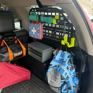 May include: A black metal MOLLE storage panel mounted in a vehicle cargo area, featuring bright neon yellow hooks holding gear. The panel displays a red and blue first aid kit, a blue and black Rawlings backpack, and a grey metal tool box.