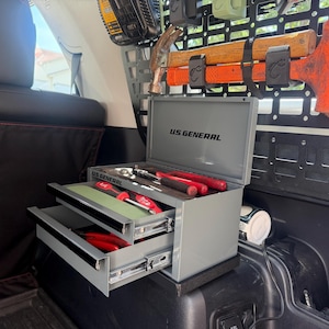 May include: A gray U.S. General metal tool chest with two open drawers containing various hand tools, positioned in a vehicle cargo area. The lid is open, displaying the brand name, and a black molle panel with a hammer and mallet is mounted behind it.