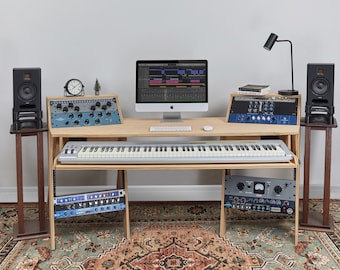 May include: A wooden music production desk with a keyboard, a computer monitor, and audio equipment. The desk has two shelves for storing equipment. The desk is set up in a home studio with a rug on the floor.