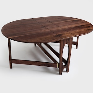 May include: A dark brown, oval-shaped wooden table with a folding design. The table has a smooth surface and is supported by angled legs with a unique cutout design. The table is approximately 60 inches long when fully extended.