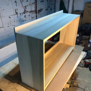 May include: A light wood shelf unit with a simple design. The shelf is made of unfinished wood and has a rectangular shape. The shelf is sitting on a piece of cardboard.