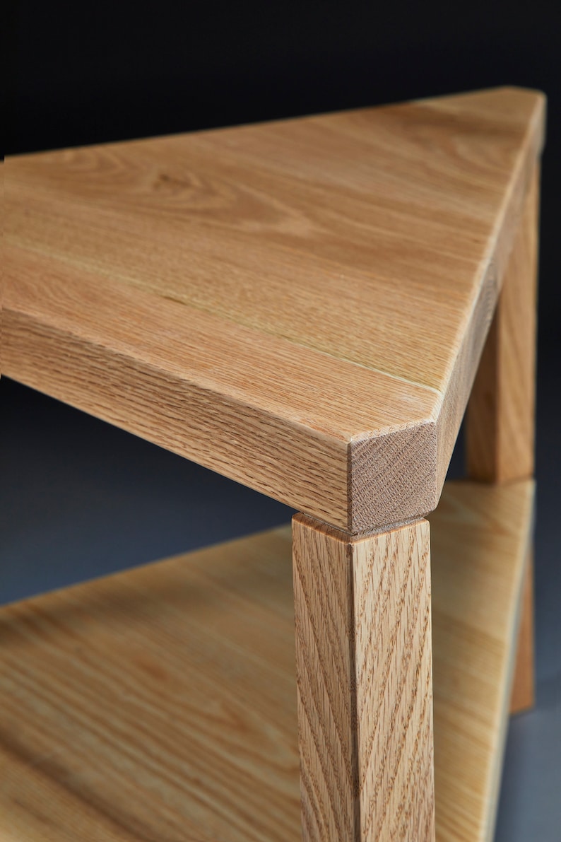 Pu&ograve; includere: Un tavolino triangolare in legno con finitura marrone chiaro. Il tavolo ha uno scaffale e una superficie superiore. La venatura del legno &egrave; visibile.