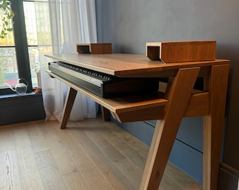 Könnte beinhalten: Ein Holzschreibtisch mit einer Tastaturablage und zwei kleinen Holzblöcken oben. Der Schreibtisch hat eine natürliche Holzoberfläche und ein modernes Design mit abgewinkelten Beinen. Die Tastatur ist schwarz. Der Schreibtisch steht in einem Raum mit Holzböden und einem Fenster.