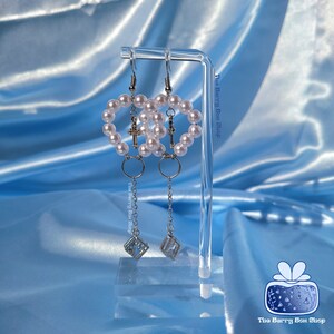 Può includere: Un paio di orecchini d'argento con un design a forma di cuore realizzato con perle bianche e croci d'argento. Gli orecchini hanno un ciondolo pendente con un cristallo trasparente.