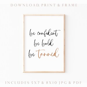 May include: A printable wall art design featuring the text "be confident be bold be tanned" in black and gold lettering on a white background. The design is framed in a light wood frame.