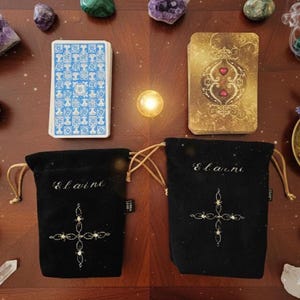 May include: An overhead shot of a wooden table with tarot cards, two black velvet drawstring bags, and crystals. The tarot cards have blue and gold designs. The bags are embroidered with a cross design and the name "Elaine".