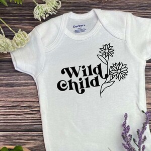 Op de afbeelding: Een witte baby romper met zwarte tekst die "Wild Child" zegt en een zwarte lijntekening van twee gestileerde bloemen.
