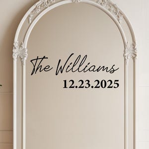 May include: A large, ornate white framed mirror with an arched top. The mirror has decorative floral accents and the text "The Williams 12.23.2025" in black script. The mirror is leaning against a neutral wall.