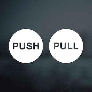 Op de afbeelding: Twee witte ronde borden met zwarte tekst op een wazige achtergrond. Het linker bord zegt "PUSH" en het rechter bord zegt "PULL".