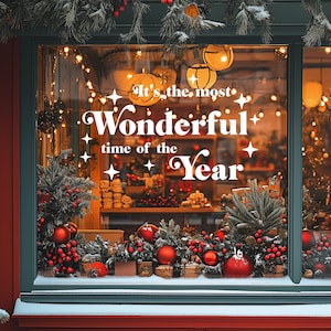 以下が含まれることがあります： クリスマスの飾り付けが施された店のショーウィンドウ。"It's the most Wonderful time of the Year"というお祝いのメッセージと、赤い実と緑の葉のディスプレイが飾られています。