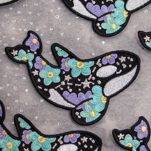 May include: Embroidered patches featuring a black orca design with white accents, adorned with teal, purple, and yellow floral patterns. The patches have a black border and are scattered across a light-colored surface.