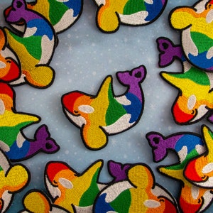 May include: A collection of rainbow-colored embroidered patches featuring orca whales. The patches are made of felt and have a white outline. The patches are perfect for adding a touch of whimsy to clothing, bags, or other accessories.