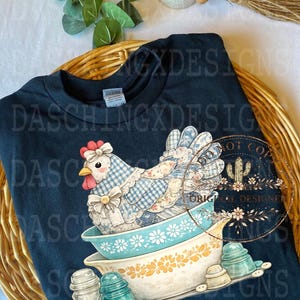 May include: Navy blue t-shirt featuring a whimsical chicken design. The chicken is depicted in a patchwork style, sitting in a vintage bowl. Additional elements include glass insulators and buttons, creating a rustic, country aesthetic.