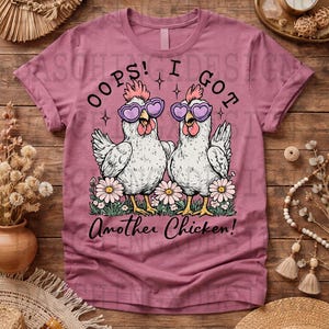 May include: A mauve t-shirt featuring a graphic of two chickens wearing heart-shaped sunglasses, standing in a bed of flowers. The text "OOPS! I GOT Another Chicken!" is printed above and below the chickens.