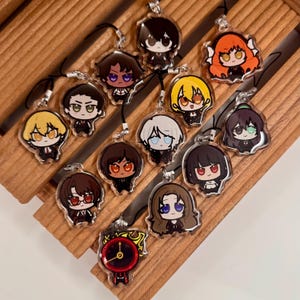 Könnte beinhalten: Eine Sammlung klarer Acryl-Anhänger mit Cartoon-Charakter-Designs mit verschiedenen Haarfarben und Gesichtsausdrücken. Jeder Anhänger hat ein schwarzes Schlüsselband. Die Anhänger sind auf einer Holzoberfläche angeordnet.