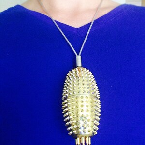 May include: A gold-coloured pendant necklace with a spiky, oval-shaped charm. The charm is covered in small, pointed studs and has a silver chain.