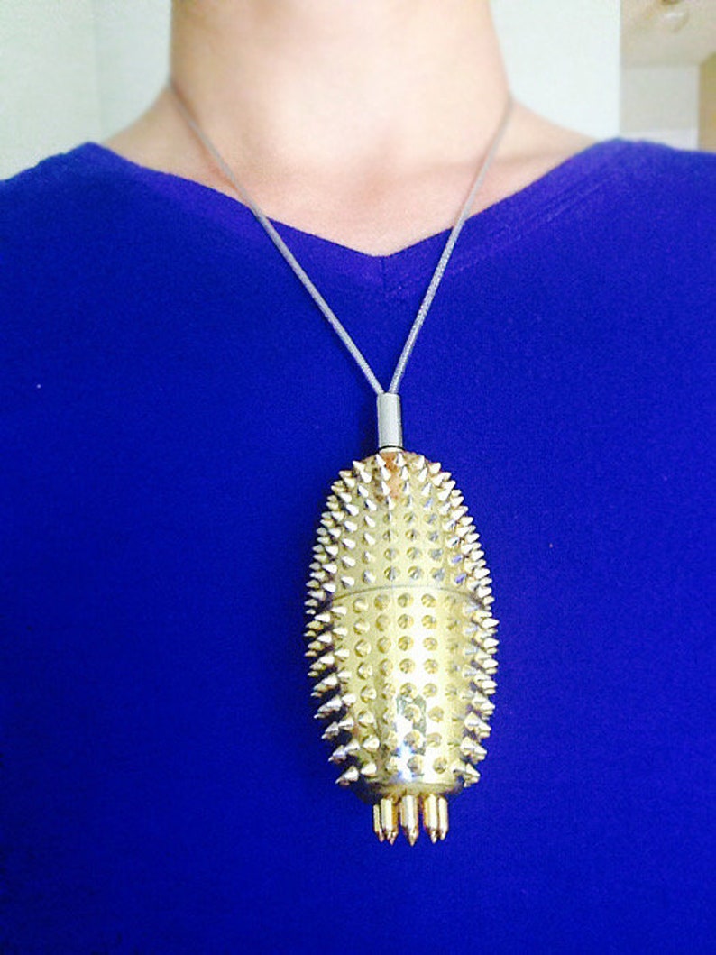 May include: A gold-coloured pendant necklace with a spiky, oval-shaped charm. The charm is covered in small, pointed studs and has a silver chain.