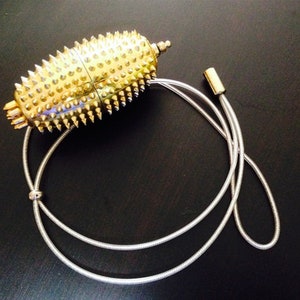 May include: A gold-coloured, spiky, oval-shaped pendant hangs from a silver chain. The pendant has a textured surface and appears to be made of metal.