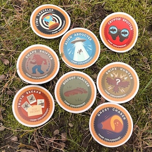 May include: A selection of circular stickers with illustrations and text such as "Time Traveller," "Alien Abductee," and "Bigfoot Tracker." Each sticker displays a unique design and the text "Camp Conjuredust."