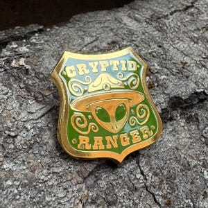 Puede incluir: Pin de metal dorado con un diseño de esmalte verde. El pin presenta un extraterrestre con un sombrero de guardabosques y el texto "Cryptid Ranger".