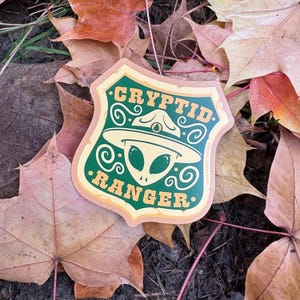May include: A green and brown sticker with a cartoon alien wearing a ranger hat. The sticker says "Cryptid Ranger" in a yellow font.