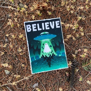Könnte beinhalten: Eine grüne, schwarze und weiße Illustration eines Geistes, der von einem UFO entführt wird. Das UFO schwebt über einem Friedhof mit Grabsteinen, auf denen "RIP" steht. Der Text "BELIEVE" befindet sich oben im Bild.