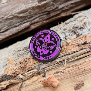 May include: A round, black and purple enamel pin with the text "CRYPTID SCOUTS OF AMERICA - TROOP 0051." The pin features a stylized emblem with a bat-like creature and stars. The pin is set against a textured wooden background.