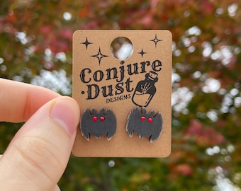 May include: A pair of black wooden bat earrings with red eyes. The earrings are on a card that says "Conjure Dust Designs" with a small bottle graphic.